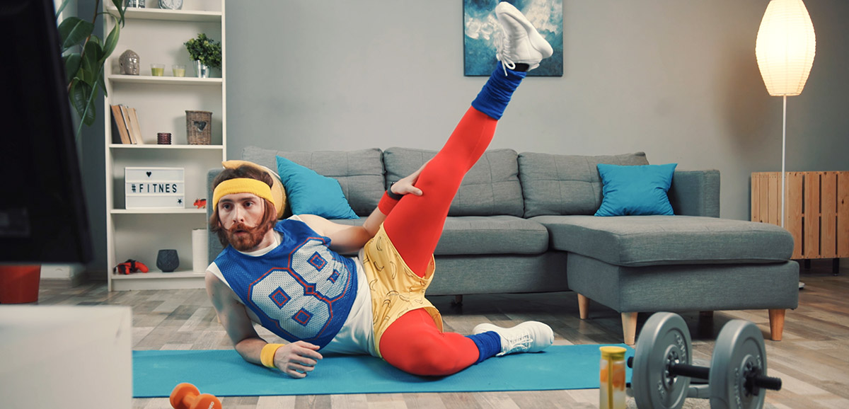 Un homme fait du sport sur un tapis devant sa télévision, il est habillé avec plusieurs couches de vêtements de sport colorés