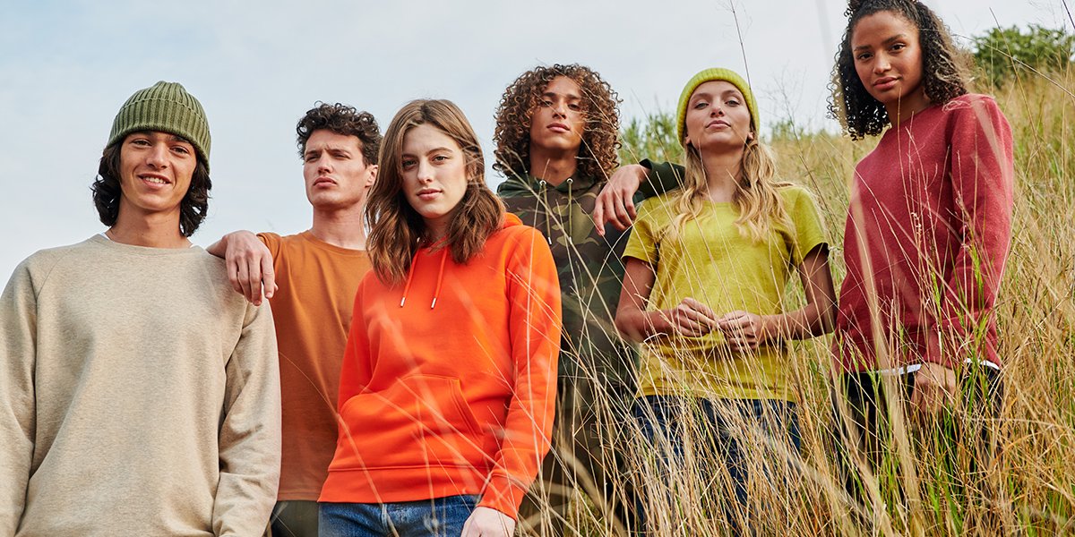 Six mannequins hommes et femmes posent dans sur une colline d'herbe en portant des sweats et t-shirts colorés de la marque Stanley/Stella